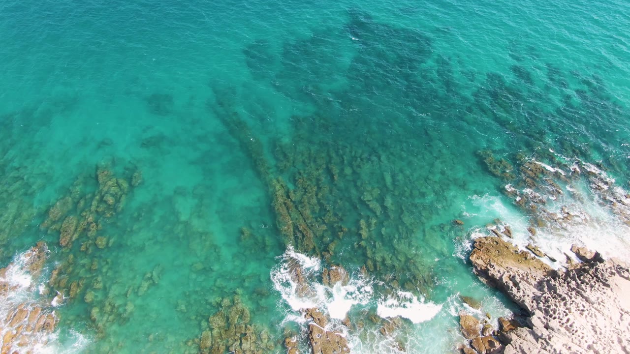Top-down Punta Gorda Beach view with a rocky outcrop near the lower frame, blue-green water, streaks of foam, and no visible people.