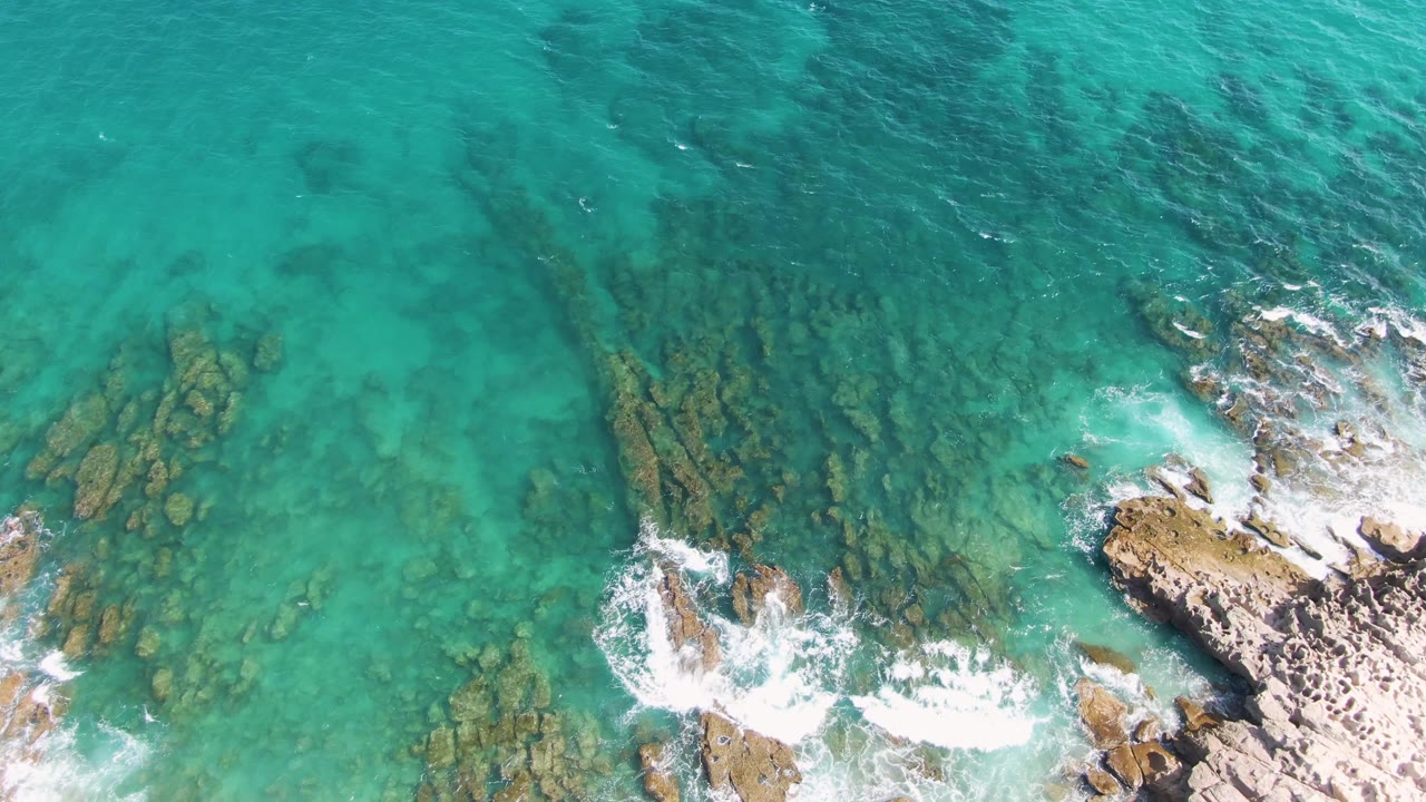 Overhead Punta Gorda Beach frame with darker submerged rock bands visible through turquoise water, breaking surf at the lower edge, and no visible people.