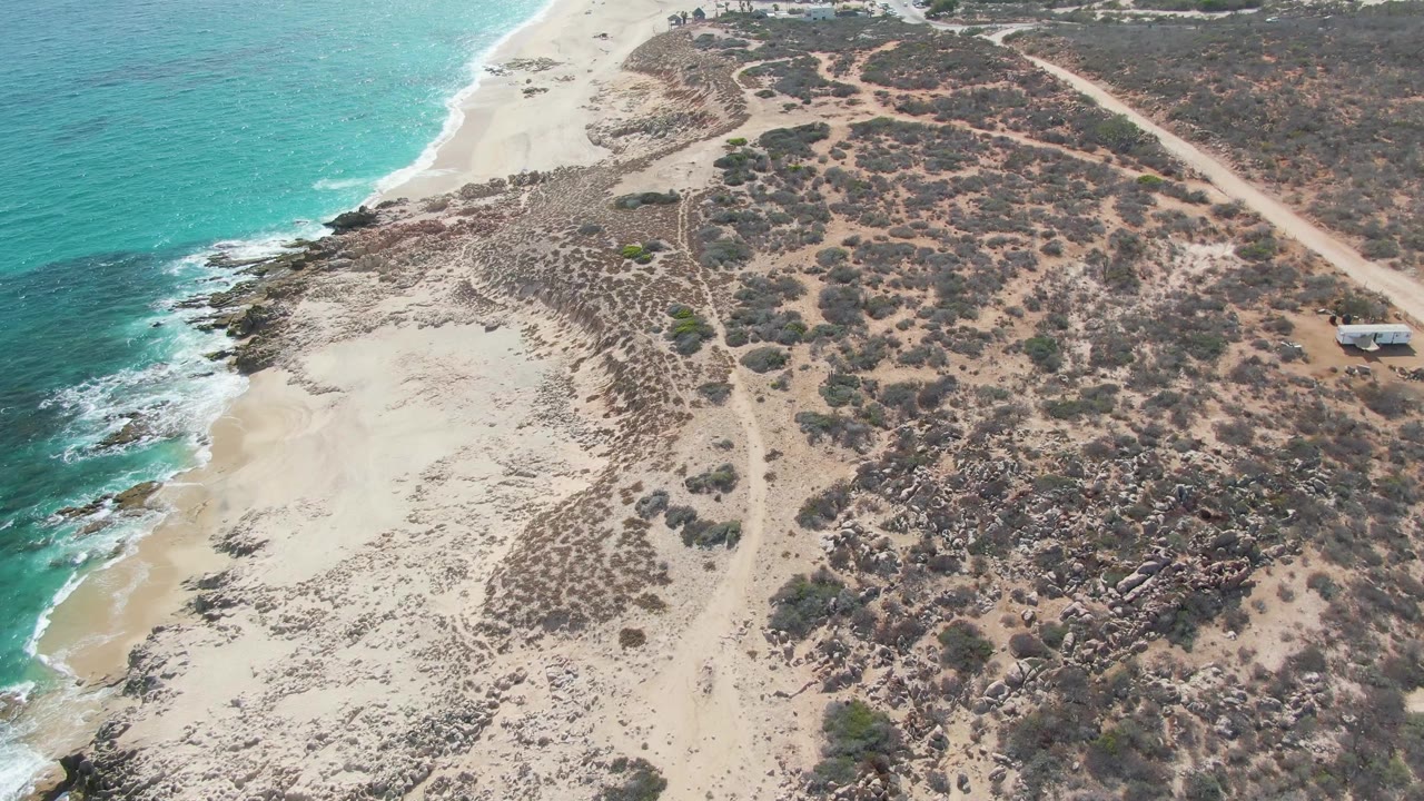 Drone view of beach dunes between turquoise water and arid scrub-covered ground.