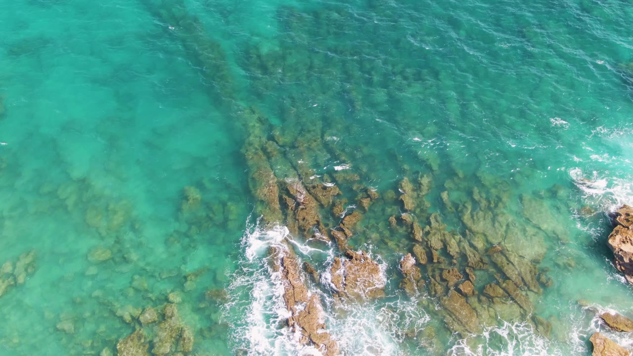 Downward drone view of turquoise water at Punta Gorda Beach with white surf curling around exposed tan rocks and darker submerged formations, with no visible people.