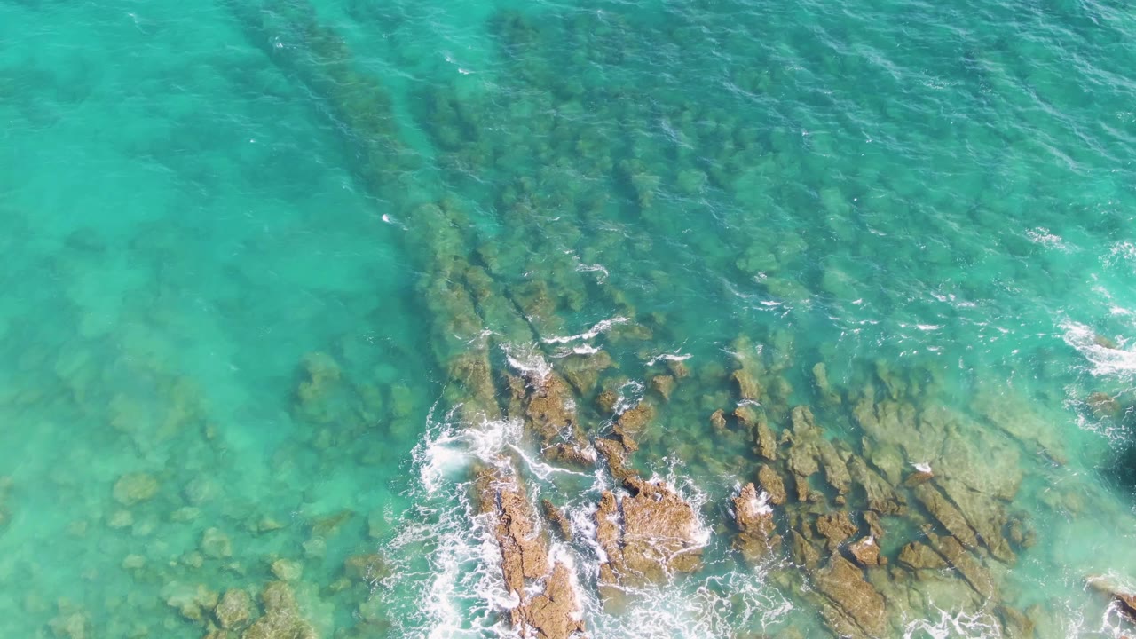 Aerial view over Punta Gorda Beach water with a broad pale turquoise area, scattered submerged rocks, whitecaps, and rocky shore detail along the bottom edge, with no visible people.
