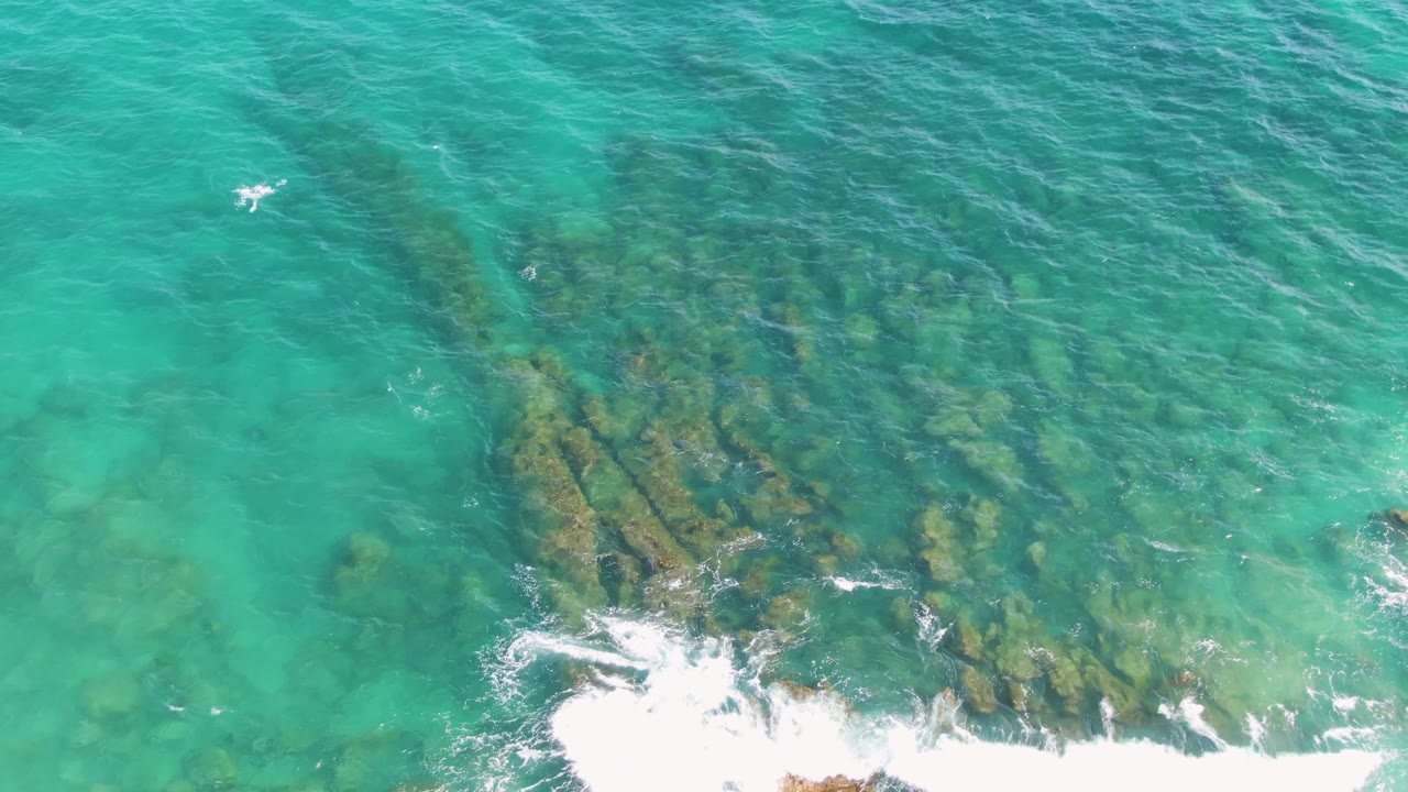 Overhead frame from Punta Gorda Beach with darker rock ridges visible under green-blue water and a line of white foam spreading along the lower portion, with no visible people.