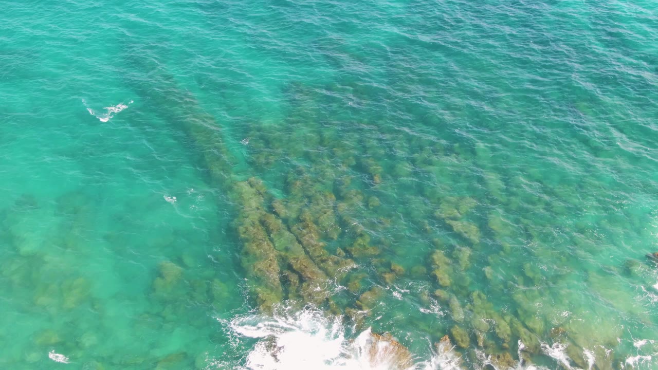 Aerial view at Punta Gorda Beach, Baja California Sur, where turquoise water reveals a clustered rocky seabed and white surf breaks along the lower right, with no visible people.