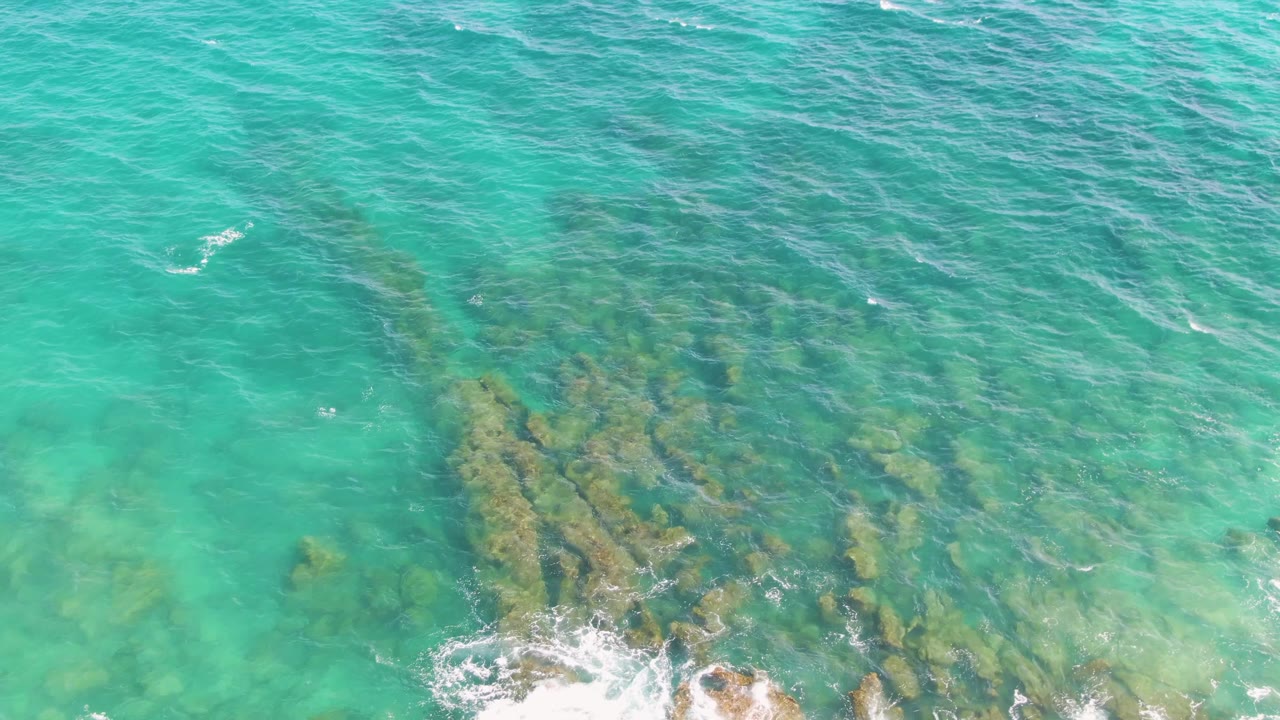Overhead Punta Gorda Beach frame showing transparent turquoise sea, uneven submerged rocks, and bright white foam pushing up from the bottom edge, with no visible people.