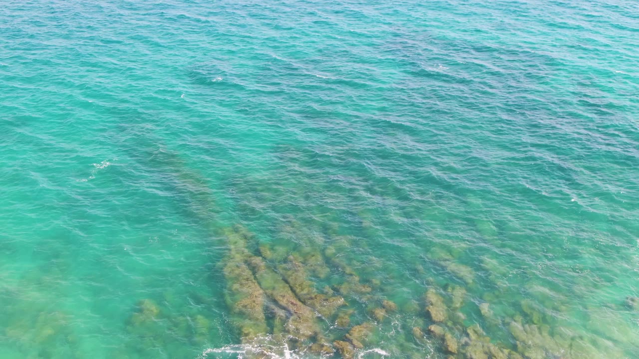At Punta Gorda Beach in Baja California Sur, clear turquoise water covers submerged rock shelves while a small line of foam reaches the lower edge, with no visible people.