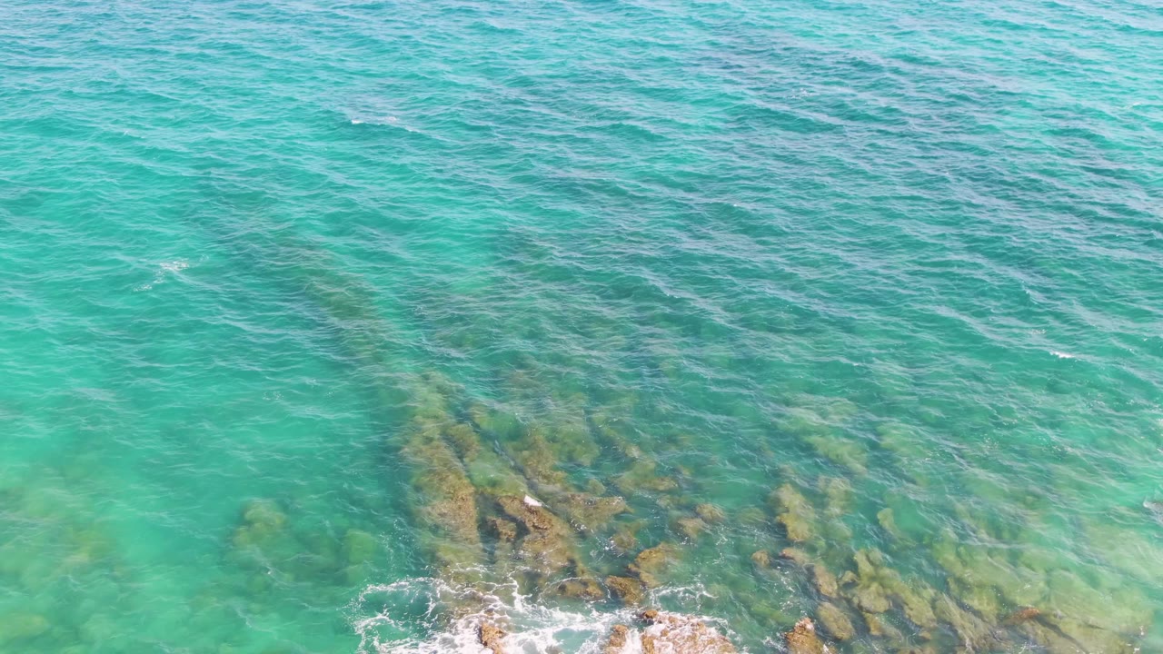 At Punta Gorda Beach in Baja California Sur, green-blue ocean water reveals rocks below the surface as a small wave begins to break near the lower center, with no visible people.