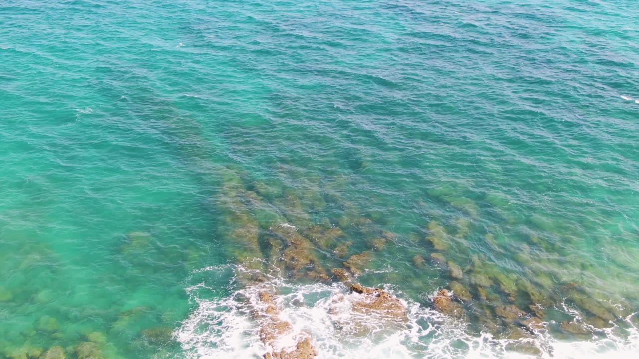 Turquoise water and submerged rocks with foamy surf spreading along the lower frame.