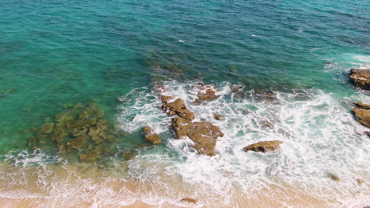 Aerial frame at Punta Gorda Beach where foamy waves break over rocks and the sandy beach begins to enter the lower edge, with no visible people.