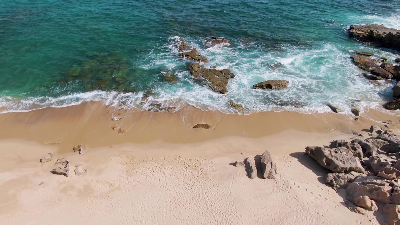 Aerial frame at Punta Gorda Beach in Baja California Sur showing turquoise water, white foam around rock outcrops, pale sand below, and no visible people.
