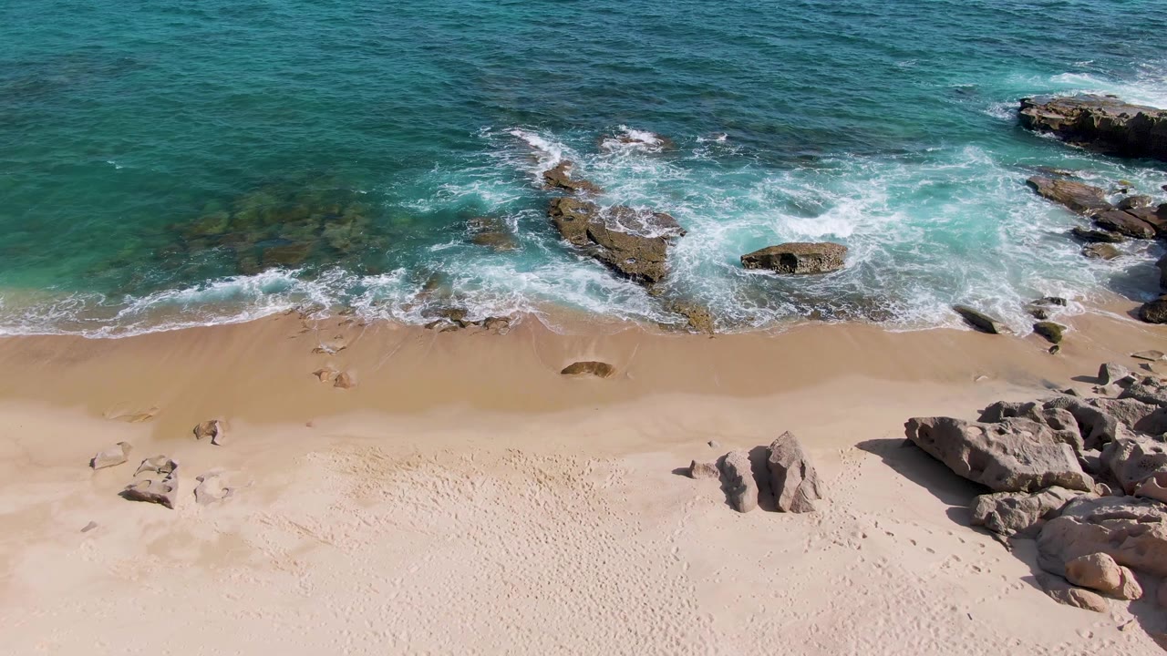 At 0:02, foam wraps the central rock cluster at Punta Gorda Beach, with deeper turquoise water above, pale beach below, and no visible people.