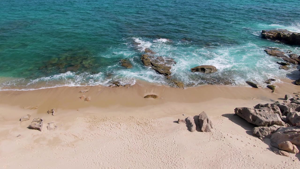 Aerial beach frame with receding foam, rocks, turquoise water, and clear sand.