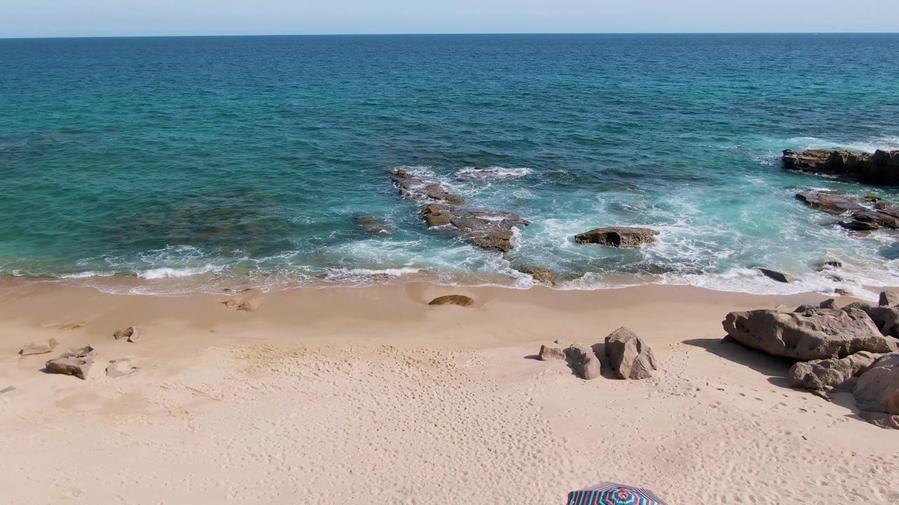 Rocky sandy beach with blue ocean water and white surf, no visible people.