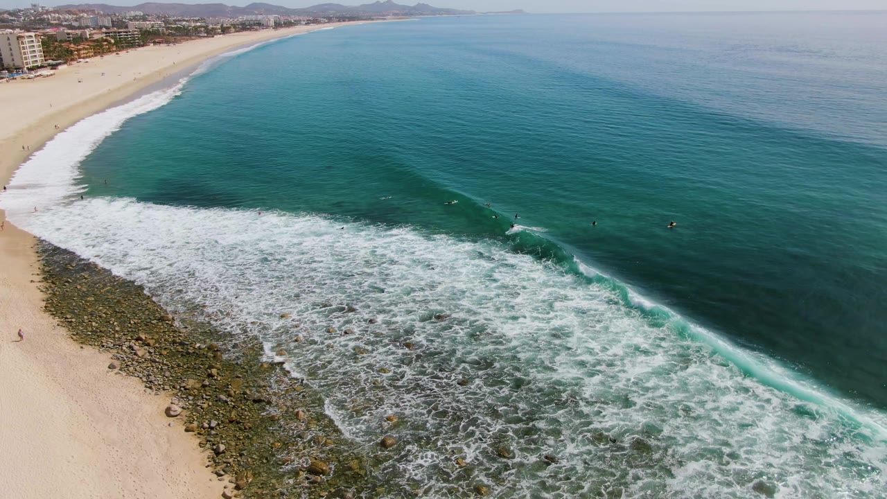 Aerial view of Punta Gorda Beach in Baja California Sur, Mexico, with white surf spreading over rocks near the sand and small distant water users offshore.
