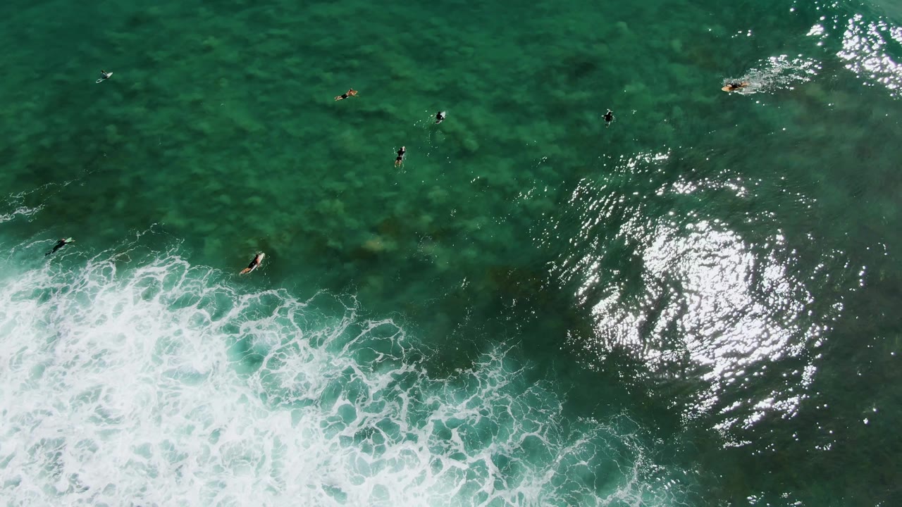 Overhead aerial frame at Punta Gorda Beach, Baja California Sur showing scattered surfers in dark and light surfboards across green-blue water, with white foam entering along the lower left edge.