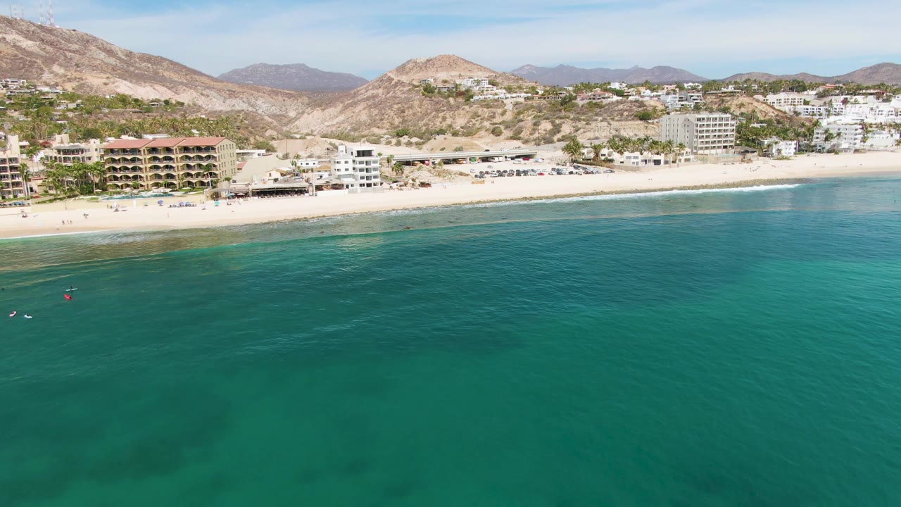 Aerial frame over Punta Gorda Beach, Baja California Sur, with turquoise water in the foreground, a pale sandy shore, beachfront buildings, dry hills, and tiny distant swimmers visible near the left water edge.