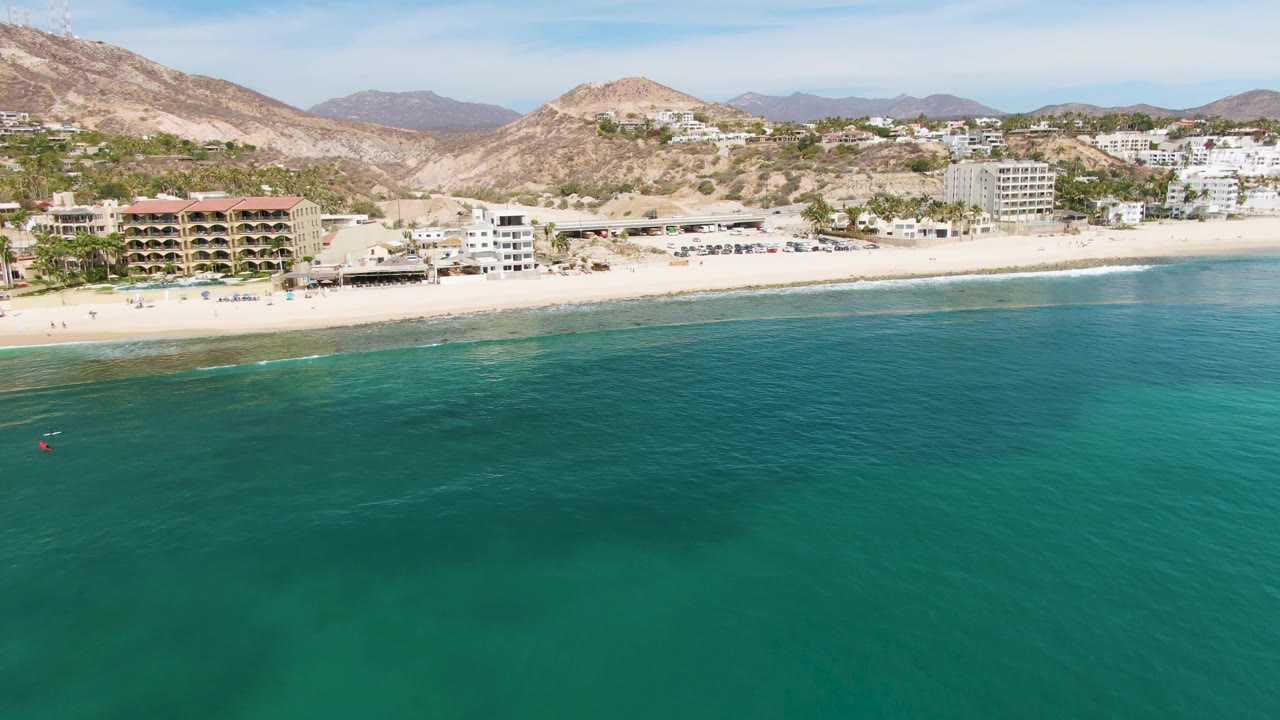 Aerial frame over Punta Gorda Beach with broad turquoise water, a narrow band of shorebreak, beachfront hotels or residences, pale sand, and dry hillside terrain under clear daylight.