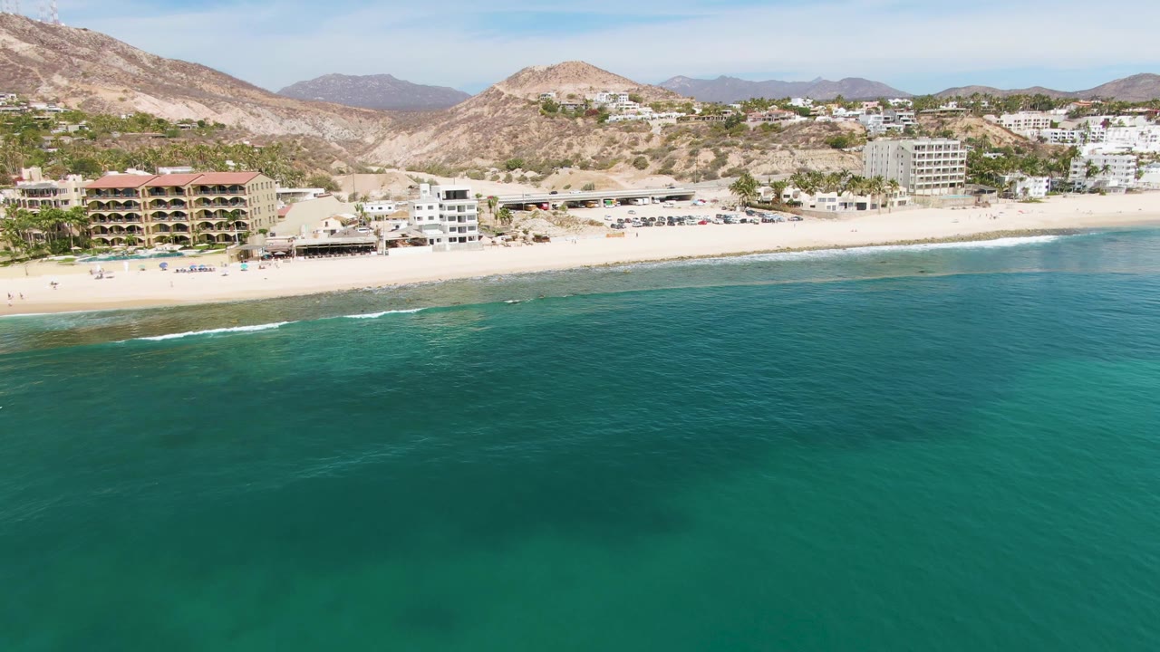Aerial view of Punta Gorda Beach showing green-blue ocean across most of the frame, light surf touching the sand, beachfront structures, and brown hills rising behind the developed shoreline.