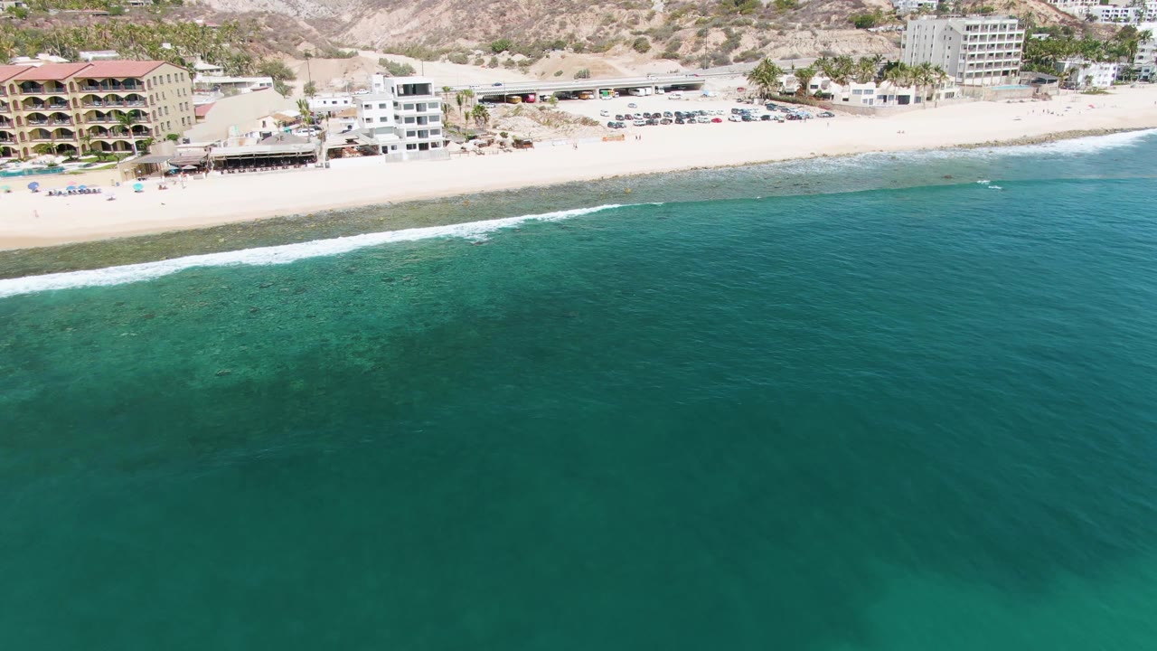 Aerial frame over Punta Gorda Beach, Baja California Sur, with turquoise water filling most of the view, sand and beachfront buildings along the top edge, and no visible people.