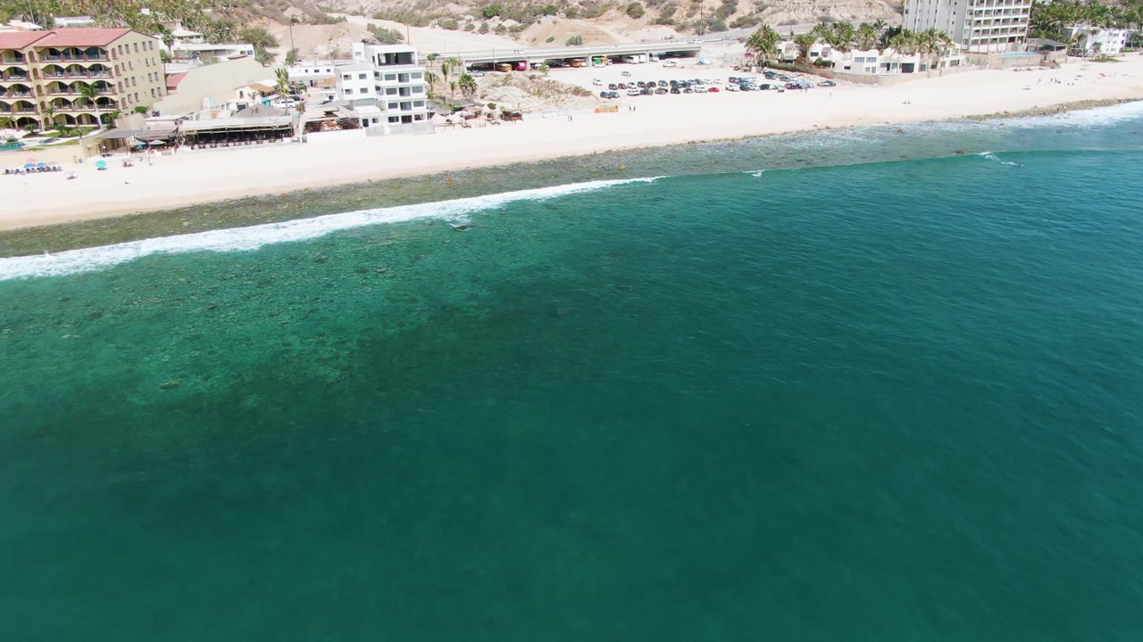 Drone view of calm turquoise water, surf, sand, and resort-like buildings.