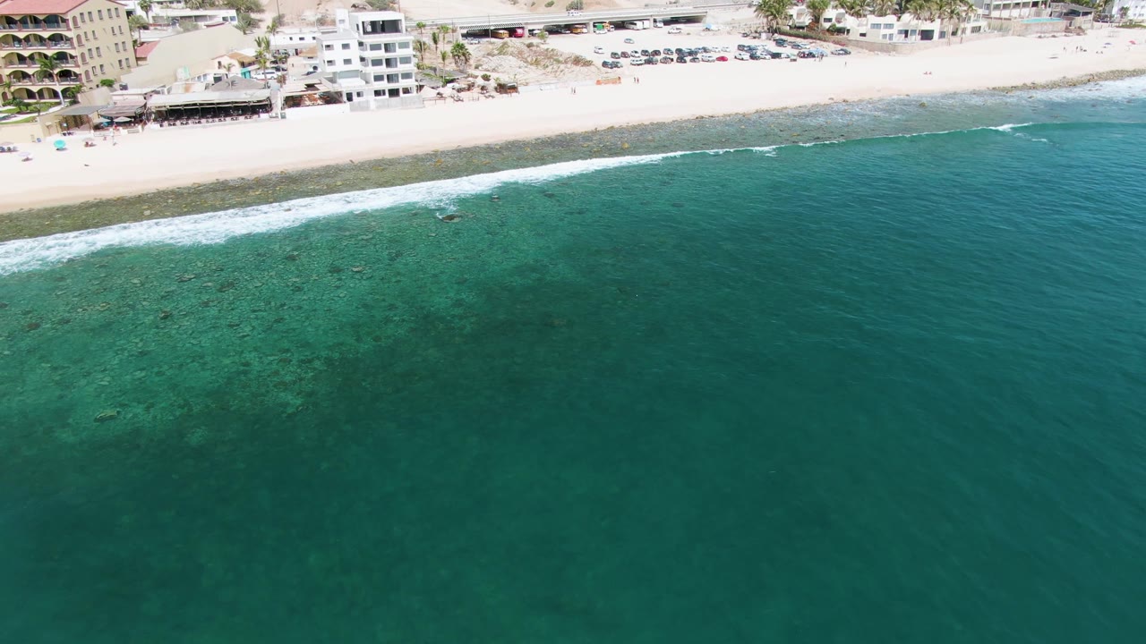 Punta Gorda Beach appears from above with green-blue water in the foreground, a bright surf band, sand, beachfront development, parked vehicles, dry hills, and no visible people.
