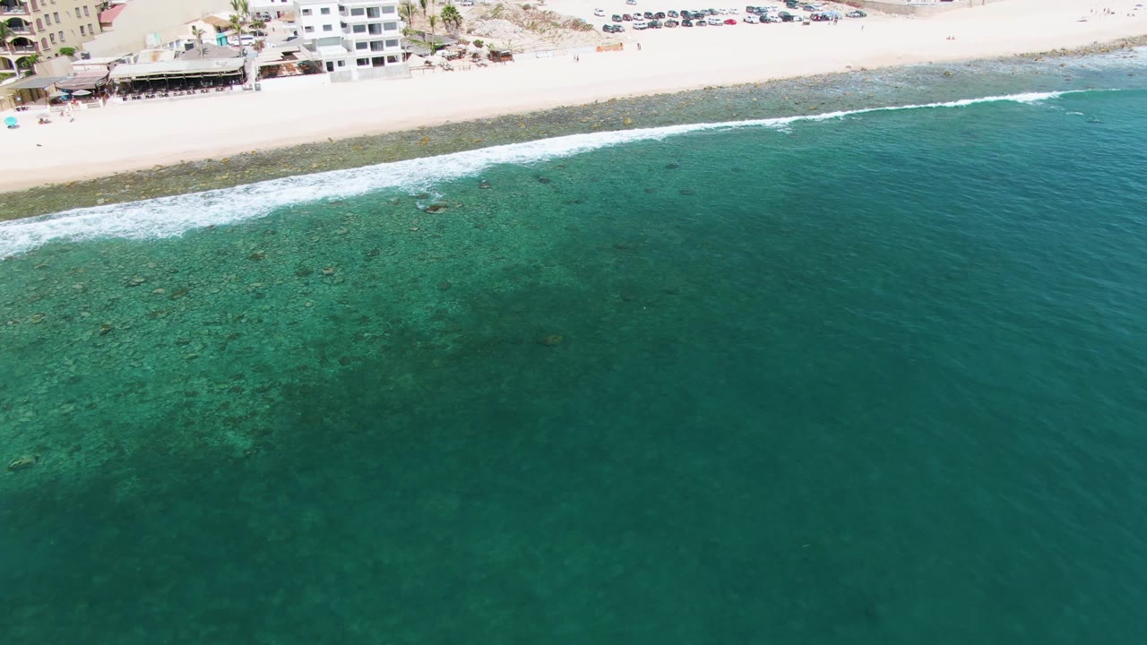 Aerial view of shallow reef-colored ocean along a sandy beach with buildings.