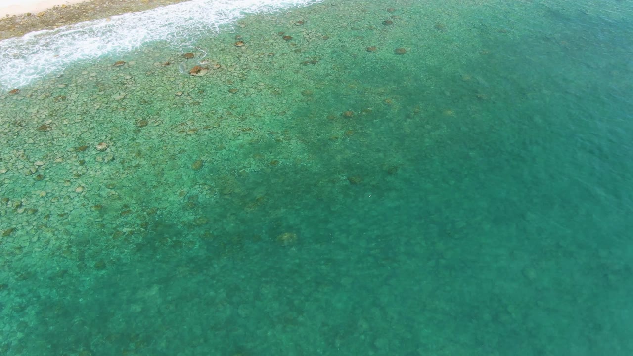 Overhead view at Punta Gorda Beach, Baja California Sur, showing white surf at the top edge and clear turquoise shallows with scattered submerged rocks, no visible people.