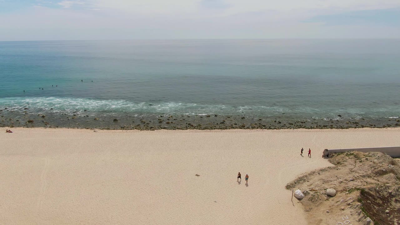 Aerial view of Punta Gorda Beach in Baja California Sur with small people walking on open sand, rocks near the shore, low surf, and a pale ocean horizon.