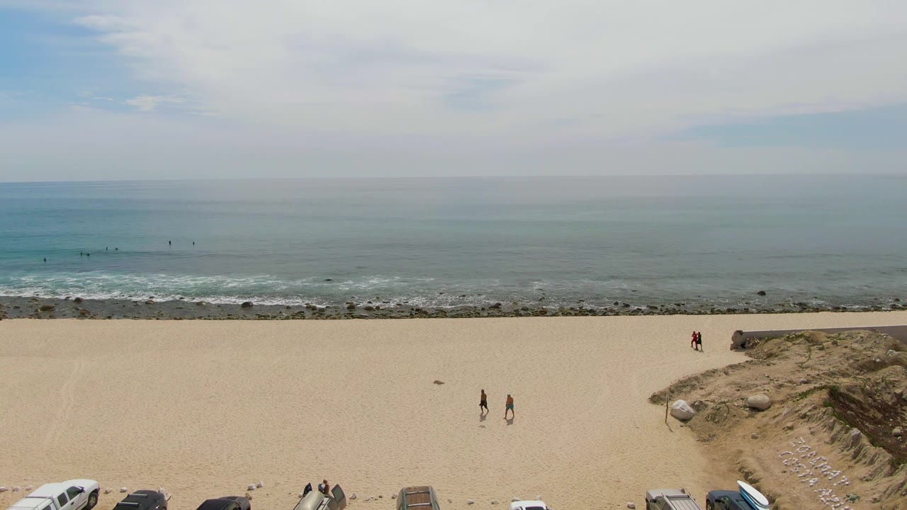 Aerial view of Punta Gorda Beach in Baja California Sur with parked vehicles along the lower edge, two small walkers on the sand, low surf, and a hazy ocean horizon.