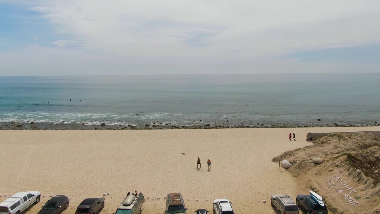 Wide aerial beach view with parked cars, people on sand, dunes, and calm ocean.