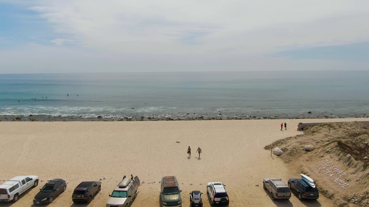 Aerial view of Punta Gorda Beach showing a fuller row of vehicles at the bottom, a pair of walkers in the center sand, two people near the dune edge, and gentle surf beyond.