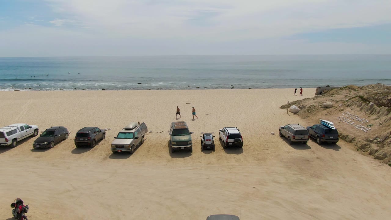 At Punta Gorda Beach in Baja California Sur, an elevated view shows parked vehicles lined along the sand, low dunes, a pale ocean horizon, and small people visible near the shore.