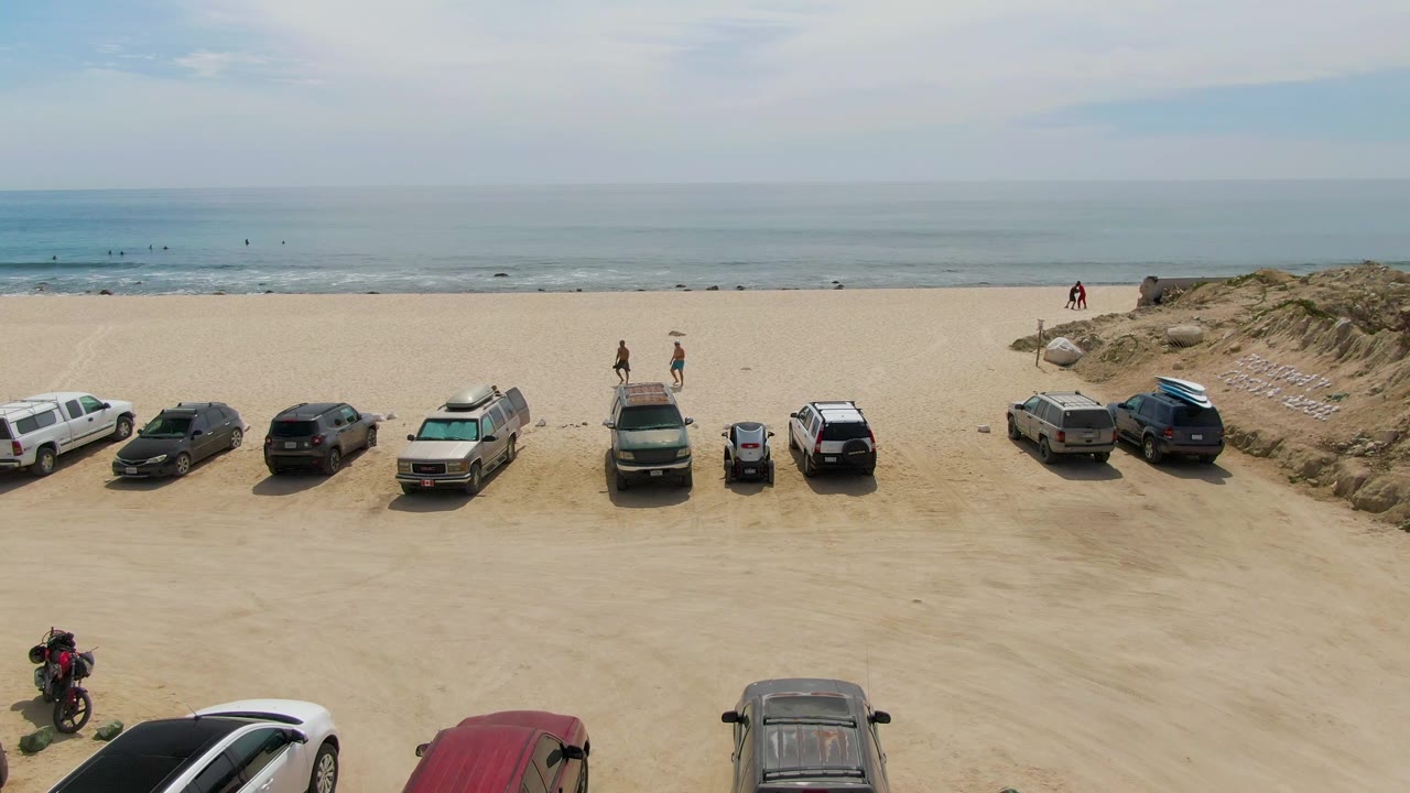 Wide beach view with parked cars, dunes, ocean, and walkers near the shore.