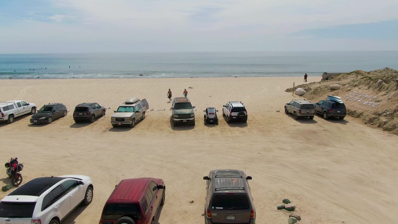 At 0:02, the aerial composition includes beach vehicles across the sand, a motorcycle near the lower edge, dunes on the right, and visible people along the Punta Gorda Beach shoreline.