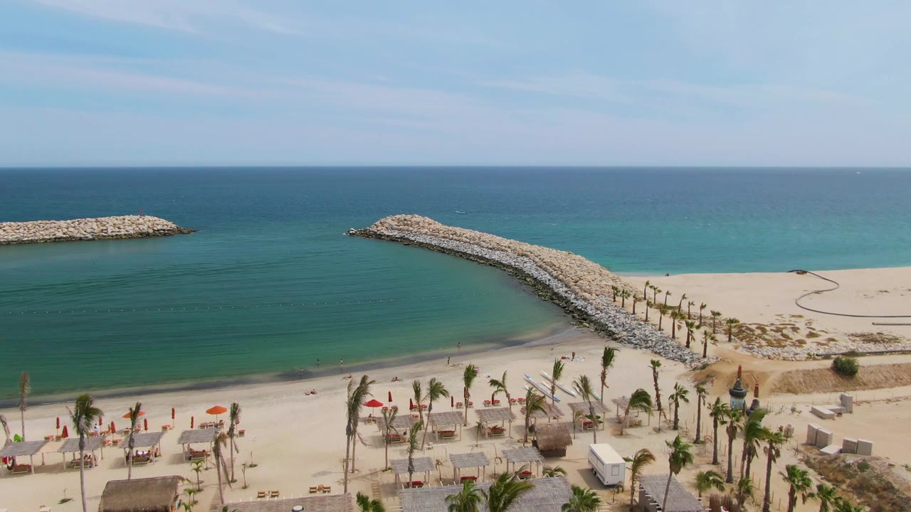 At 0:00, Puerto Los Cabos Beach appears from above with a curved stone breakwater, calm turquoise water, pale sand, palms, umbrellas, chairs, and no visible people.