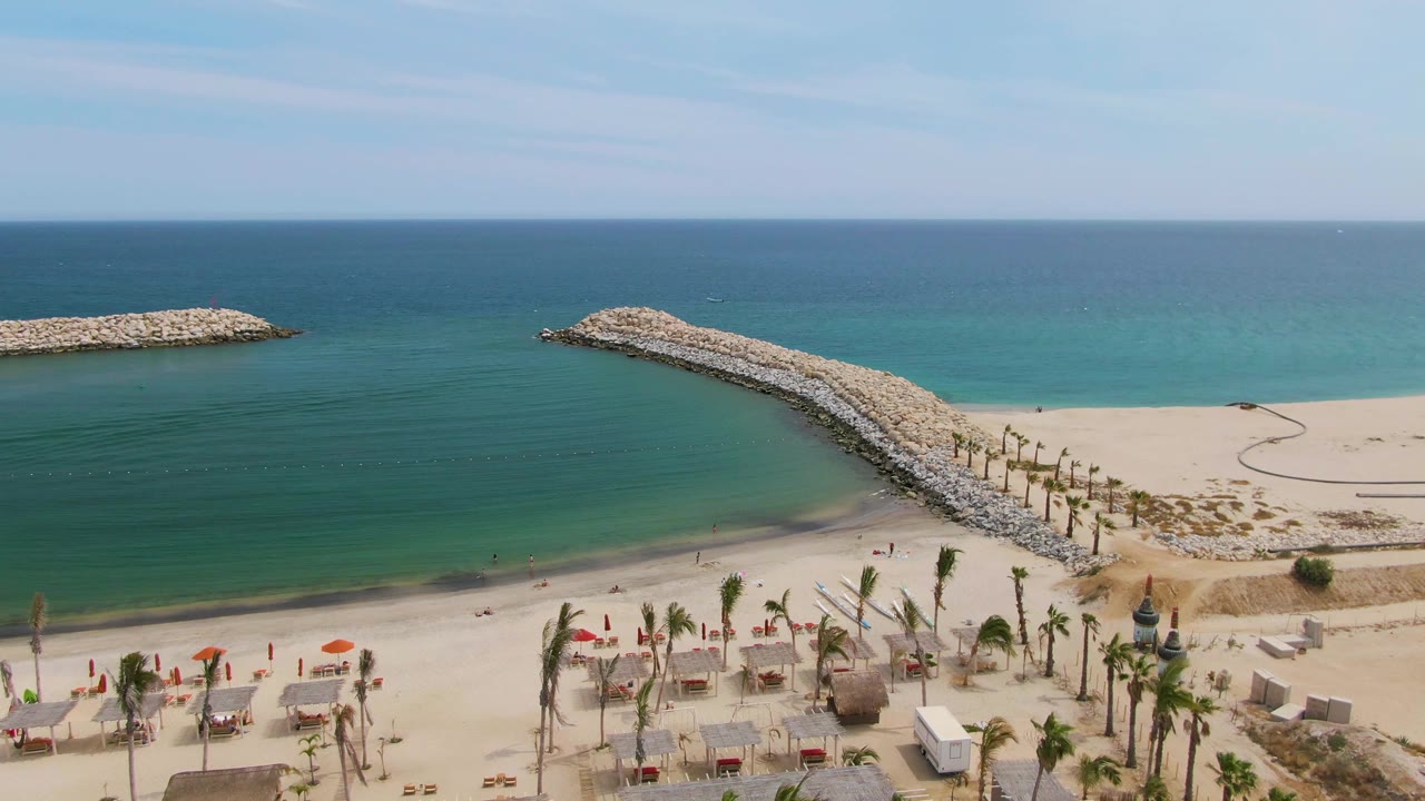 At 0:01, the curved rock jetty divides blue-green water and sand at Puerto Los Cabos Beach, with palm rows and beach seating near the lower frame and no visible people.