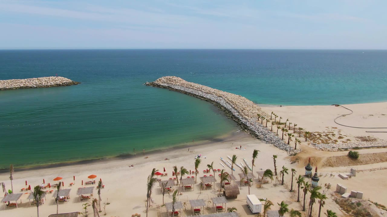 At 0:02, Puerto Los Cabos Beach shows the stone breakwater extending diagonally from the sand into calm water, with a detached rock arm offshore and no visible people.