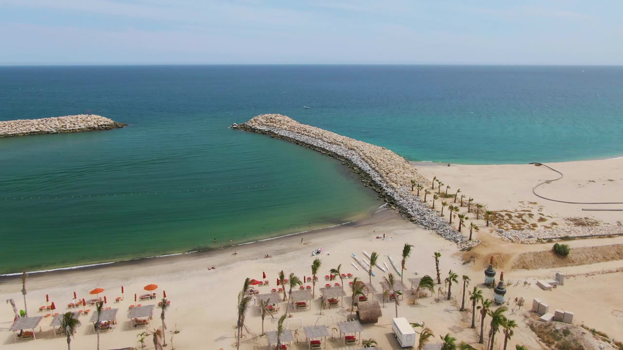 Beachfront palms and umbrellas below a curved rock breakwater