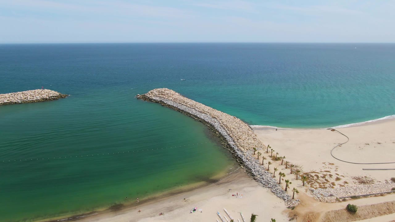 Aerial view of curved rocky breakwater beside sandy shore and clear turquoise ocean.