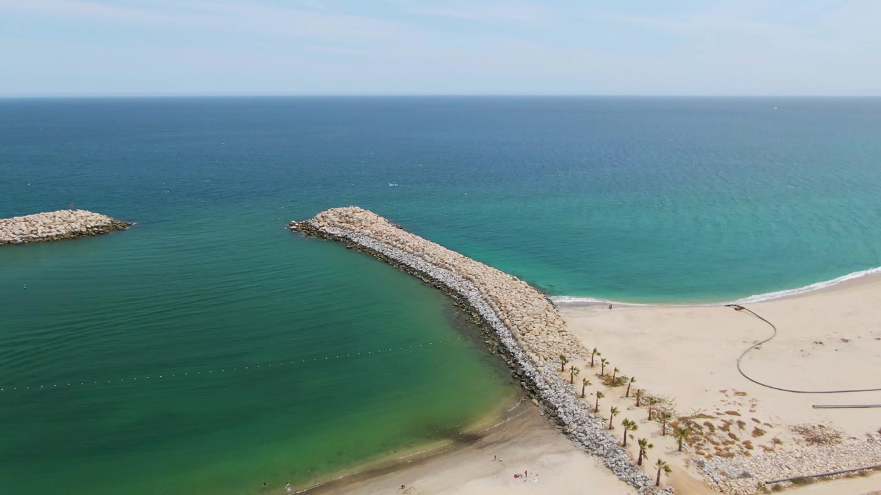 Puerto Los Cabos Beach aerial frame showing the breakwater angled from the lower right into green-blue water, with a mostly open sandy beach and a small offshore rock structure.