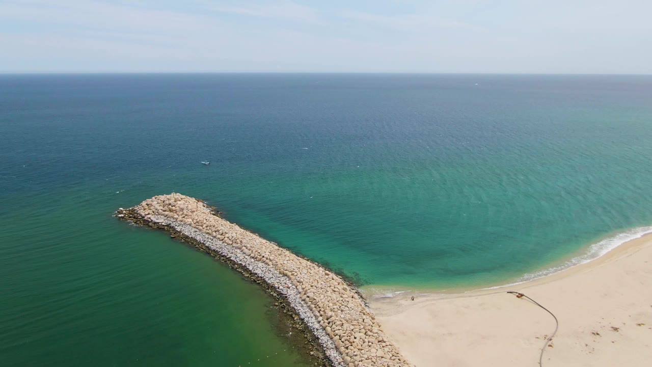Aerial view at Puerto Los Cabos Beach showing a curved rocky groyne leading from pale sand into blue-green ocean, with no visible people.