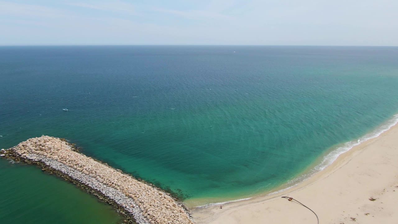 Puerto Los Cabos Beach appears from above with the groyne angled into the ocean, green-blue water around the rocks, light surf along the sand, and no visible people.