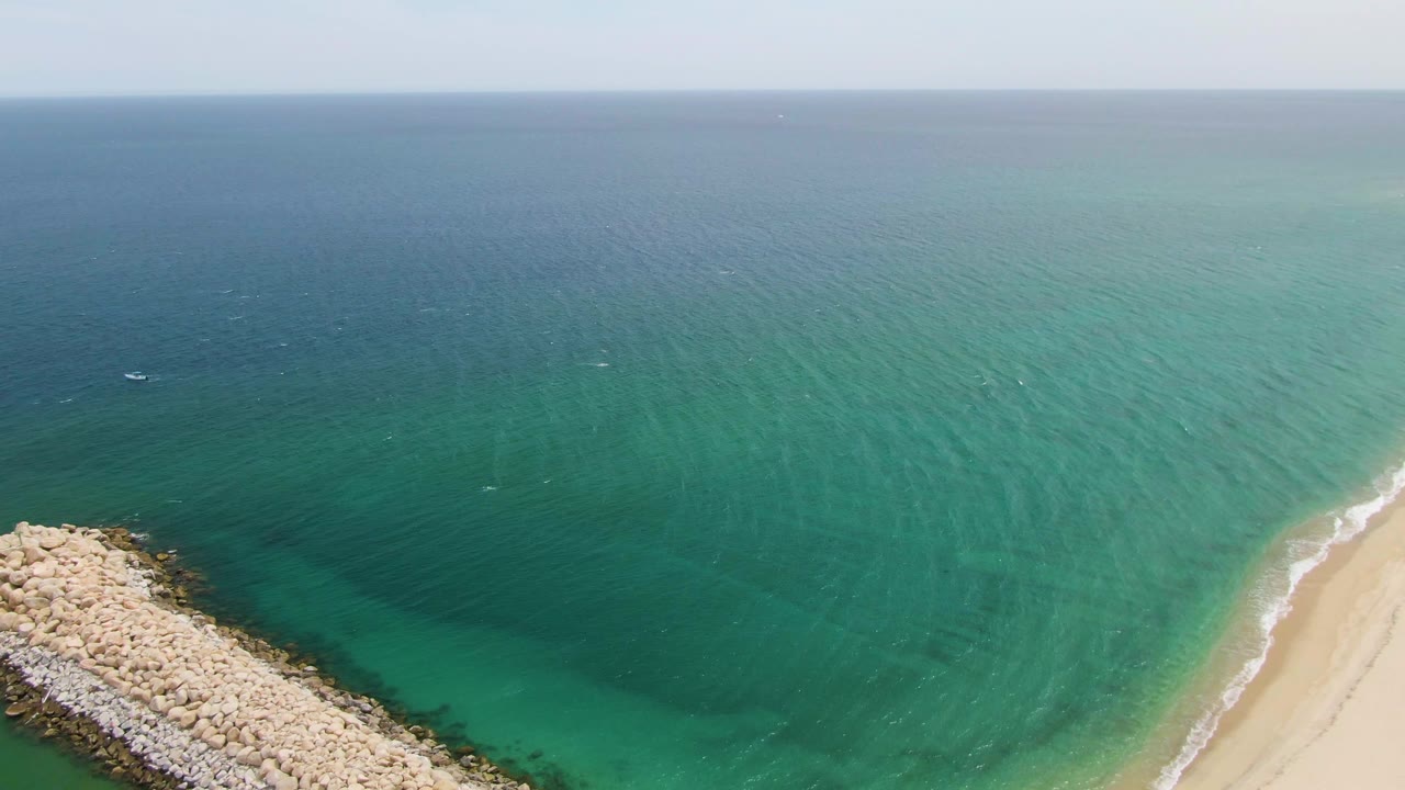 Aerial frame over Puerto Los Cabos Beach with a rocky breakwater at lower left, pale sand at lower right, turquoise water across the frame, a hazy horizon, and no visible people.