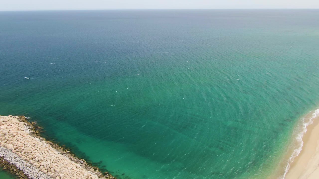 Aerial view at Puerto Los Cabos Beach with the rock breakwater centered lower frame, sand to the right, darker water beyond the shallows, faint offshore boats, and no visible people.