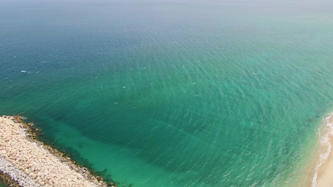 Aerial frame over Puerto Los Cabos Beach with the breakwater rising from the lower center, shallow green-blue water on both sides, pale sand at right, and no visible people.