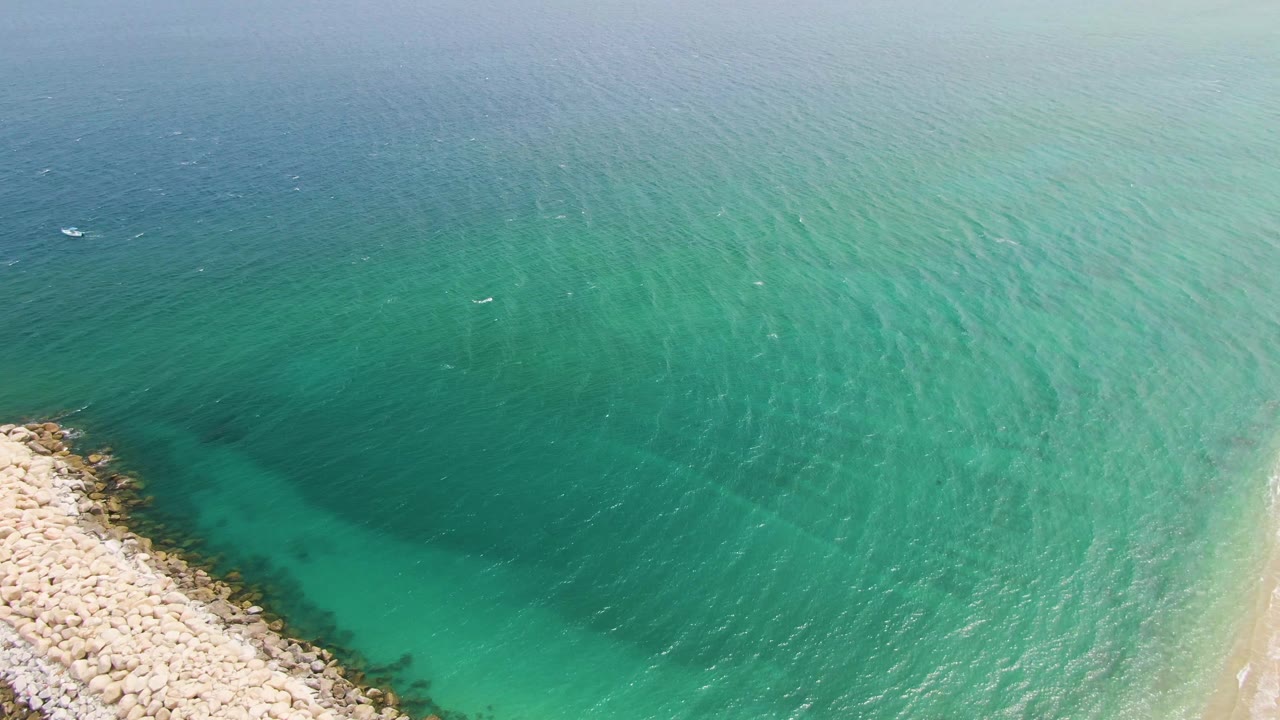 Puerto Los Cabos Beach appears from above with rocks in the lower left, a narrow strip of sand, broad turquoise water with visible ripples, small distant boats, and no visible people.