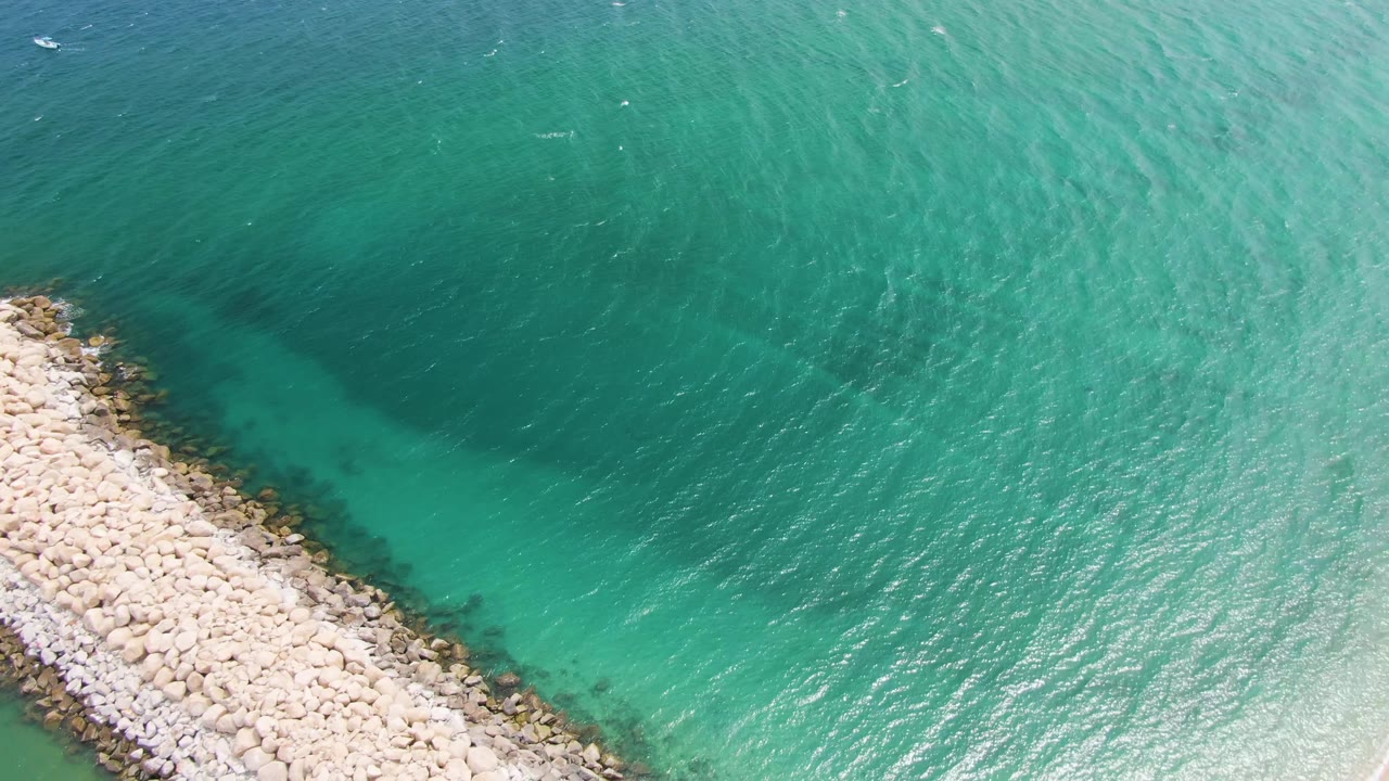 Aerial frame over Puerto Los Cabos Beach shows a pale rock breakwater entering from the lower left into clear turquoise water, with no visible people.