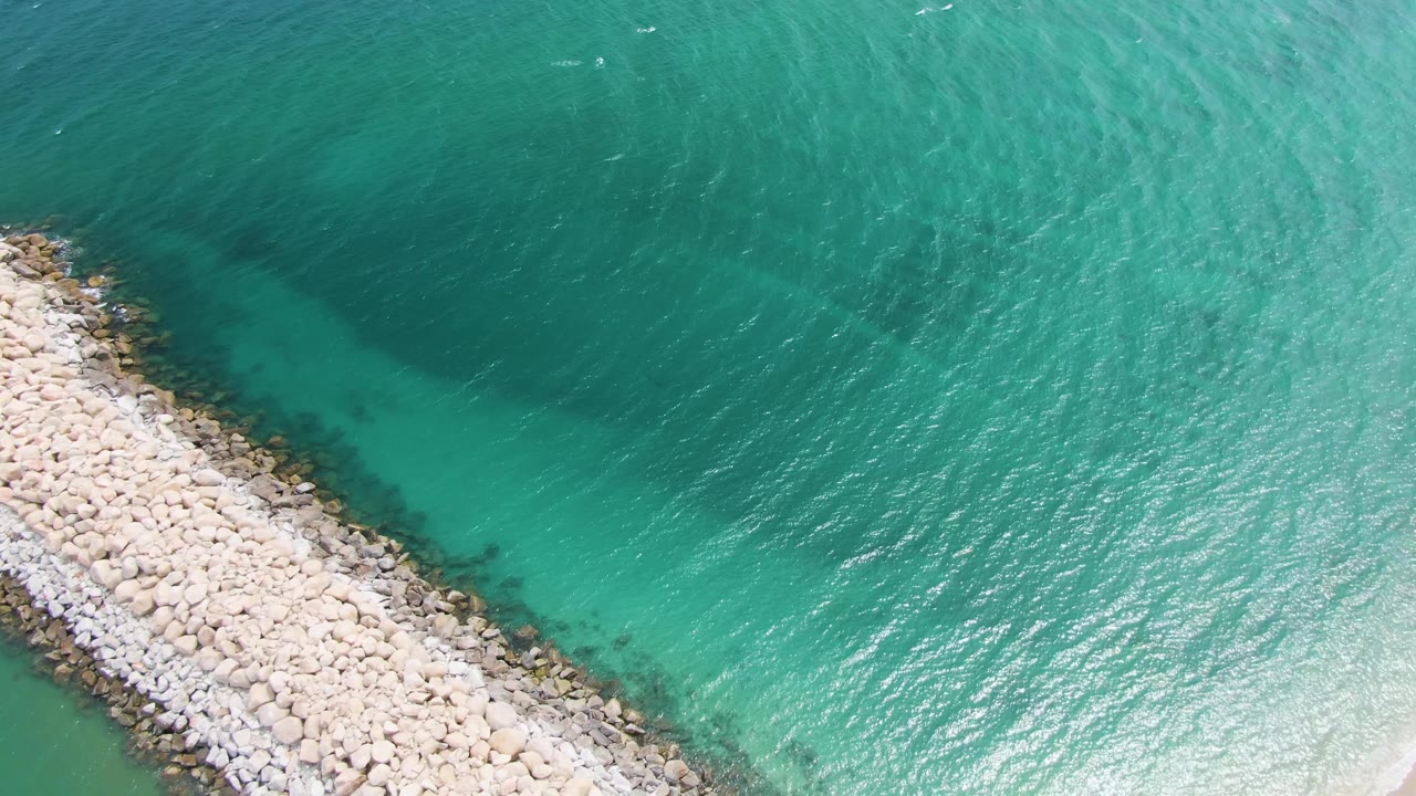 At 0:01, the Puerto Los Cabos Beach breakwater sits more centrally in the aerial view, bordered by darker shallow water and bright turquoise ripples, with no visible people.