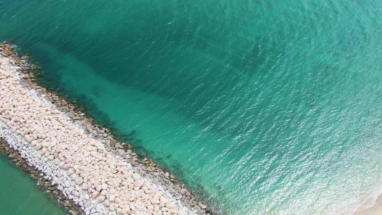 At 0:02, the aerial angle keeps the Puerto Los Cabos Beach rock barrier running diagonally through turquoise water, with brighter ripples on the right and no visible people.