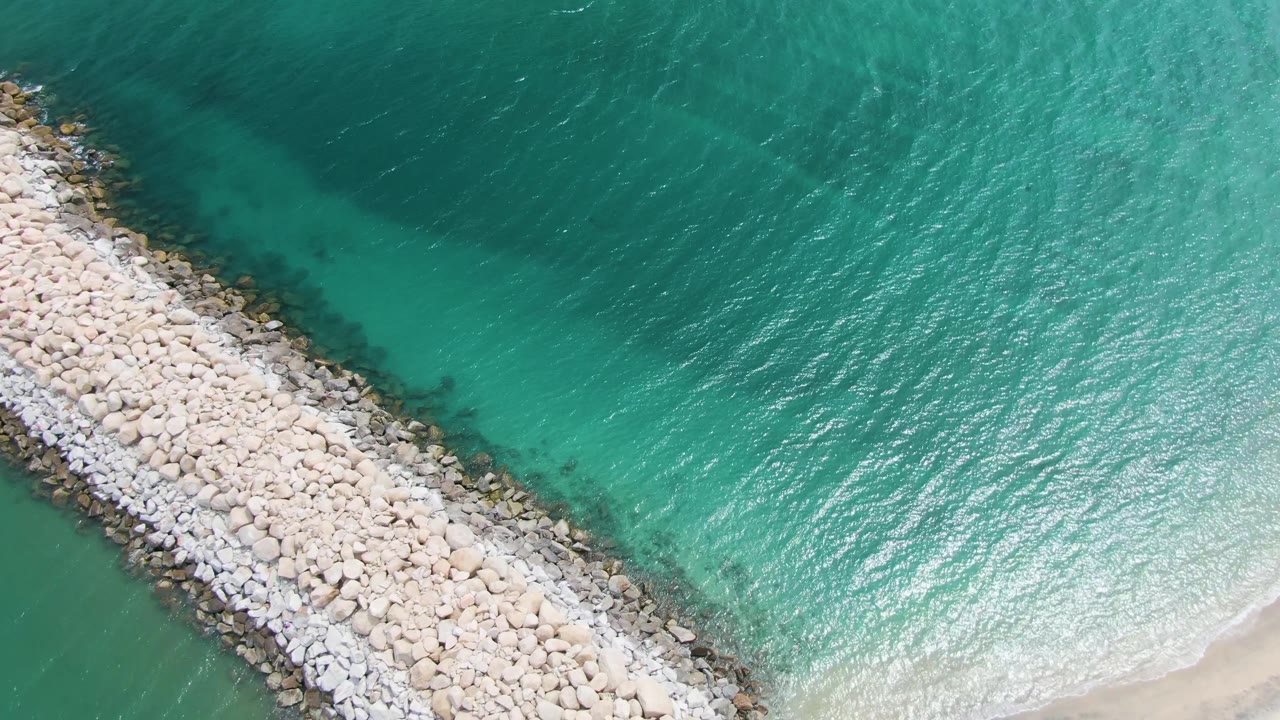 Rock breakwater beside turquoise water with sandy shore beginning to appear.