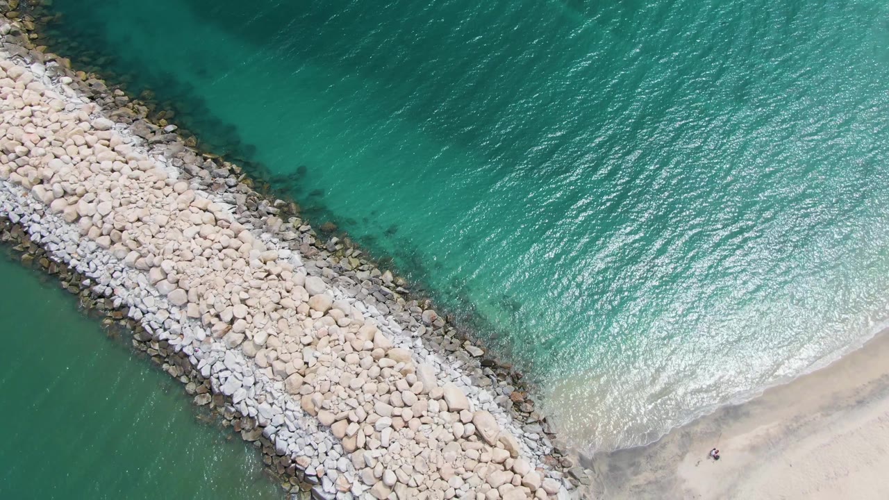 Aerial view at Puerto Los Cabos Beach showing a pale rock breakwater angled across turquoise ocean with sandy shore at the lower right and no visible people.